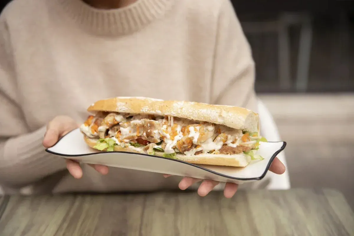 Manos sosteniendo un sándwich en un plato sobre una mesa de madera, con un fondo interior desenfocado.