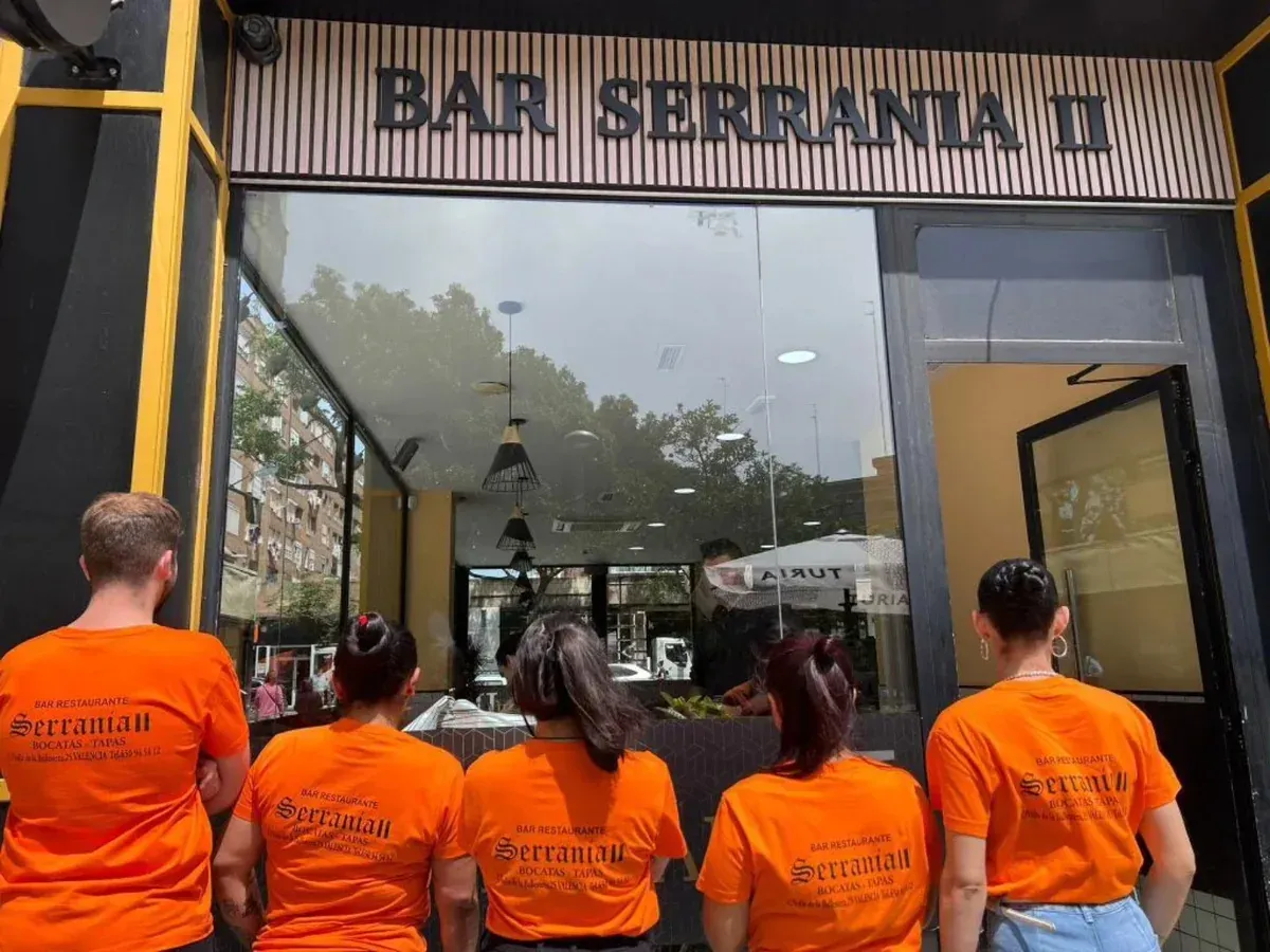 Cinco personas con camisetas naranjas están de pie frente al Bar Seranta II, mirando hacia la fachada del local.