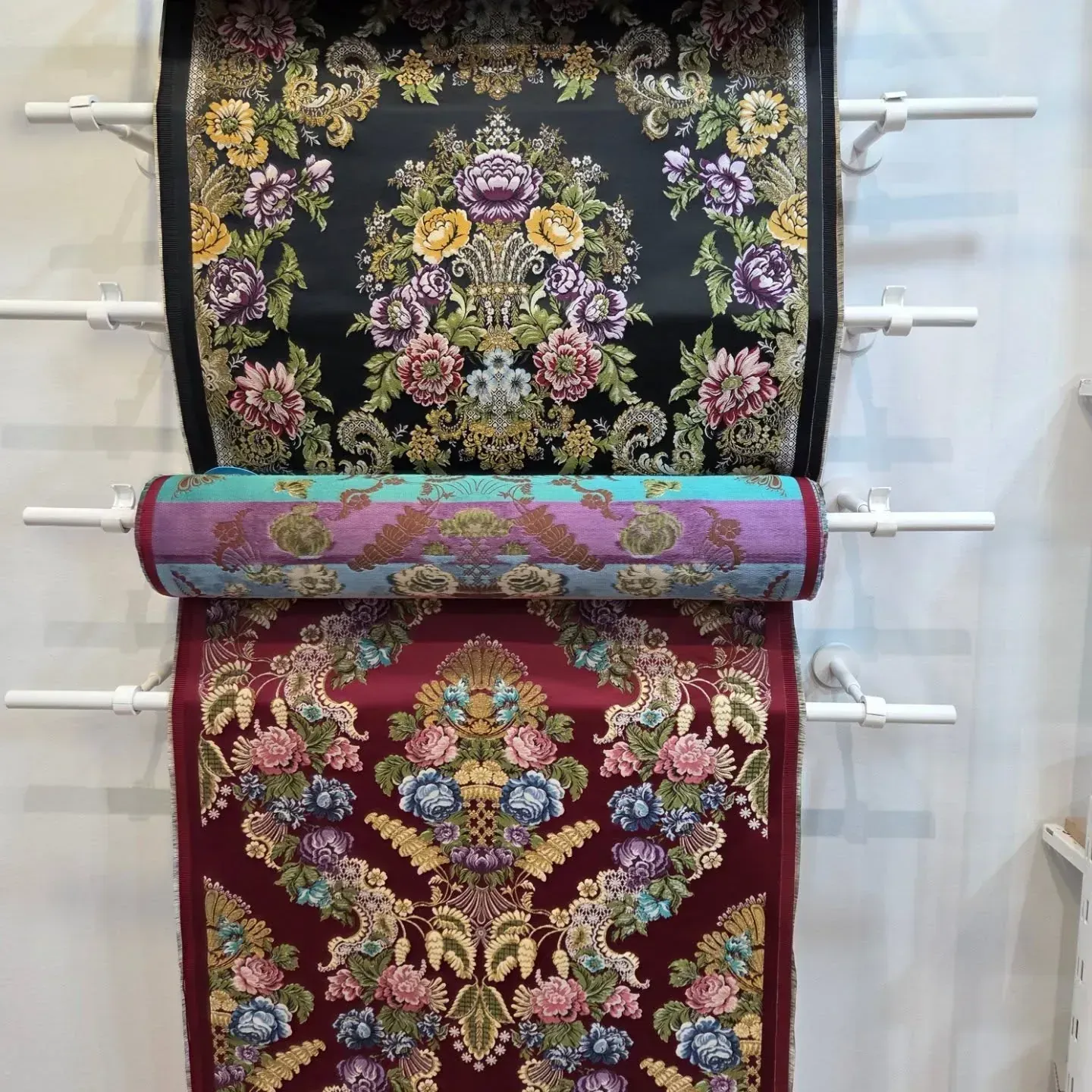 Tres rollos de tela con estampado floral en negro, verde azulado y burdeos colgados en estantes blancos.