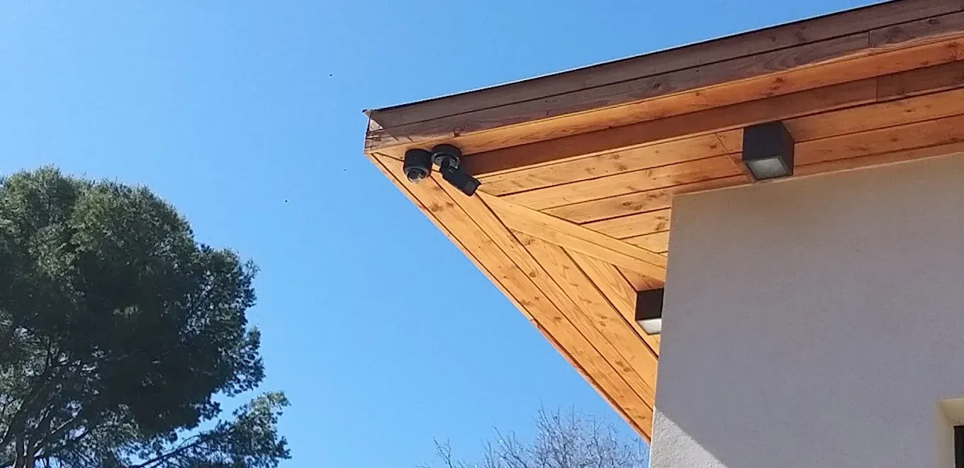 Toit d'une maison en bois sur fond de ciel bleu éclatant. Un arbre se trouve à gauche, et des luminaires carrés y sont installés.