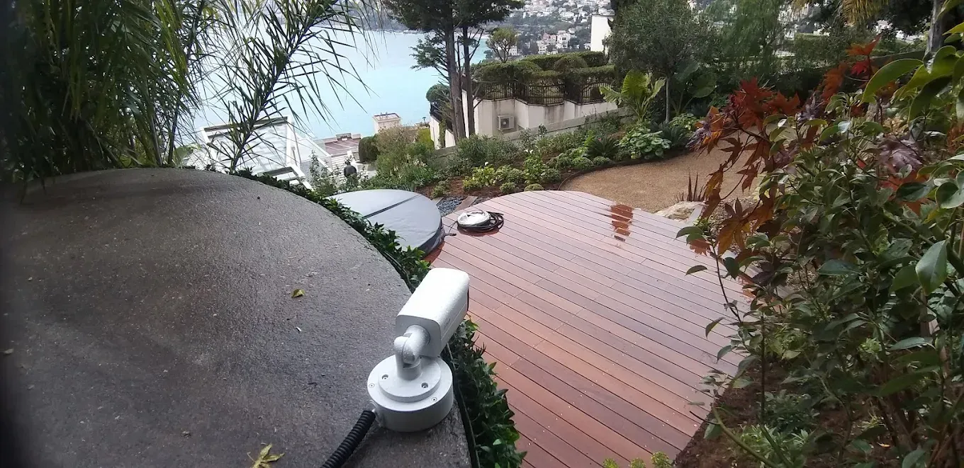 Vue d'en haut sur une terrasse en bois et un rivage avec une caméra de sécurité au premier plan.