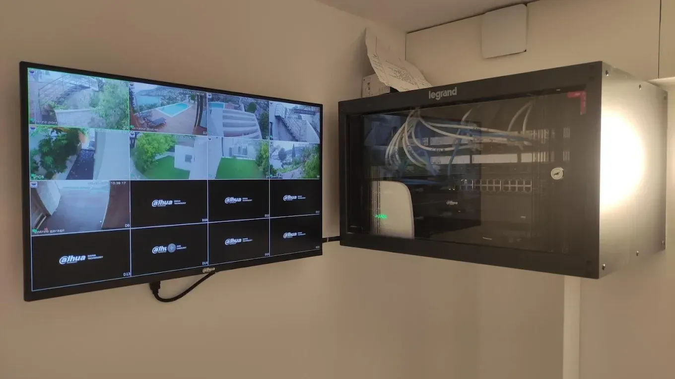 Un moniteur de sécurité mural affiche les flux de plusieurs caméras. Un rack accueille les composants électroniques, les câbles étant visibles. Murs blancs, intérieur.