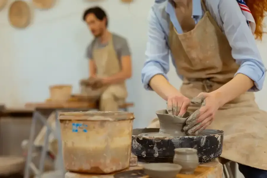 Mujer modelando arcilla en un torno de alfarero, hombre al fondo. Ambos llevan delantales en un taller.