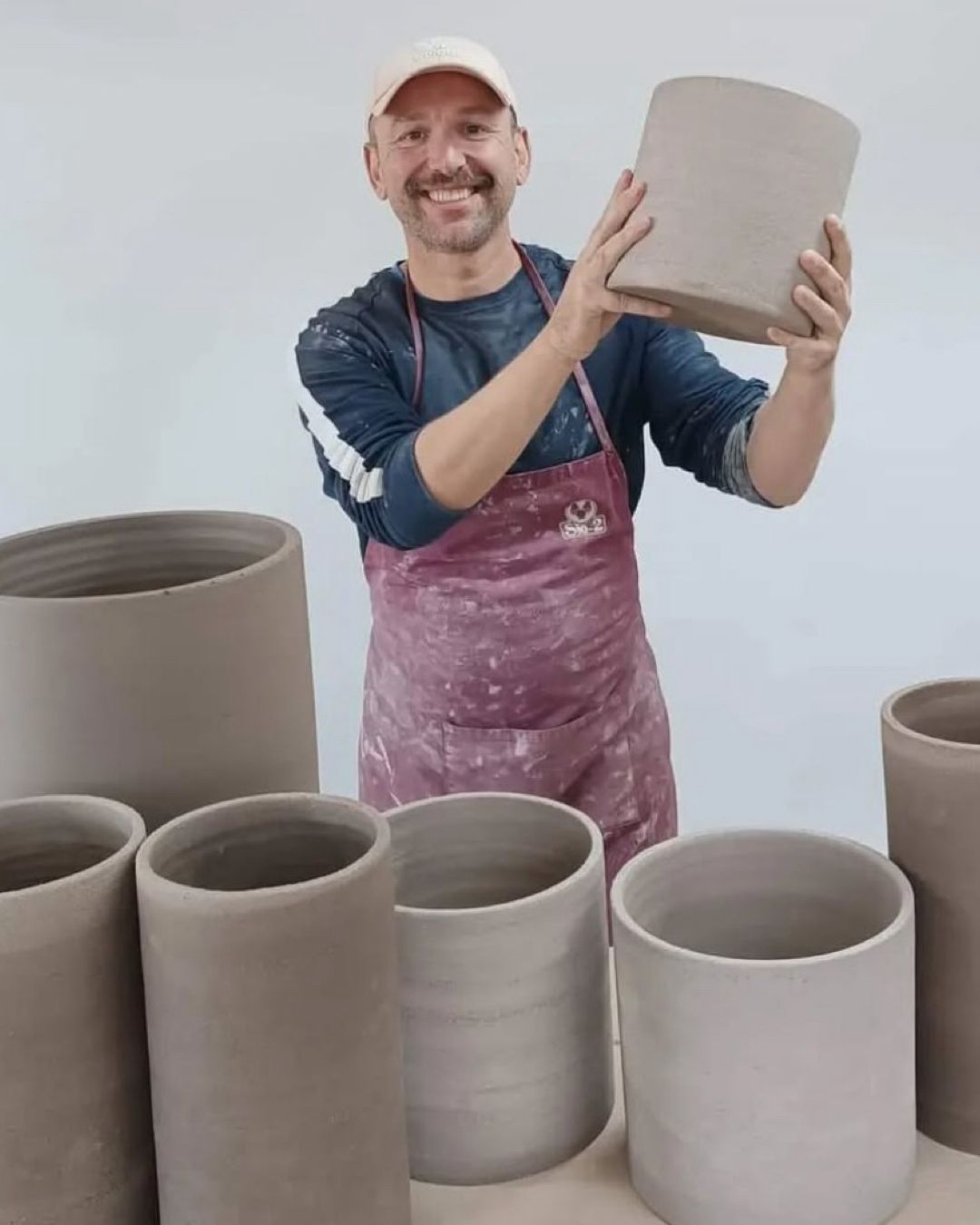 Mujer modelando arcilla en un torno de alfarero, hombre al fondo. Ambos llevan delantales en un taller.