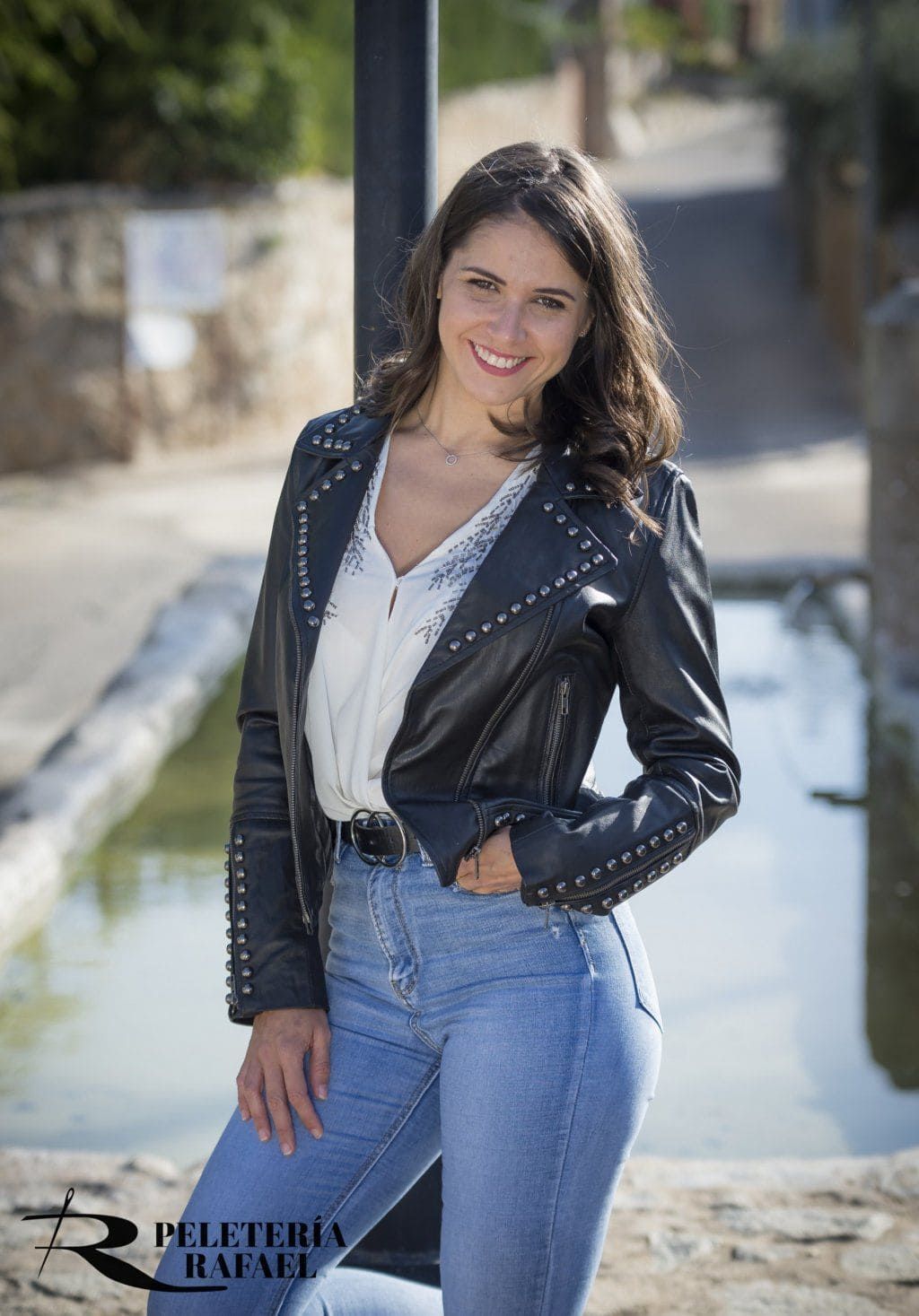 Una mujer con una chaqueta de cuero negra tachonada, una camiseta blanca y vaqueros sonríe al aire libre cerca de una fuente de agua.