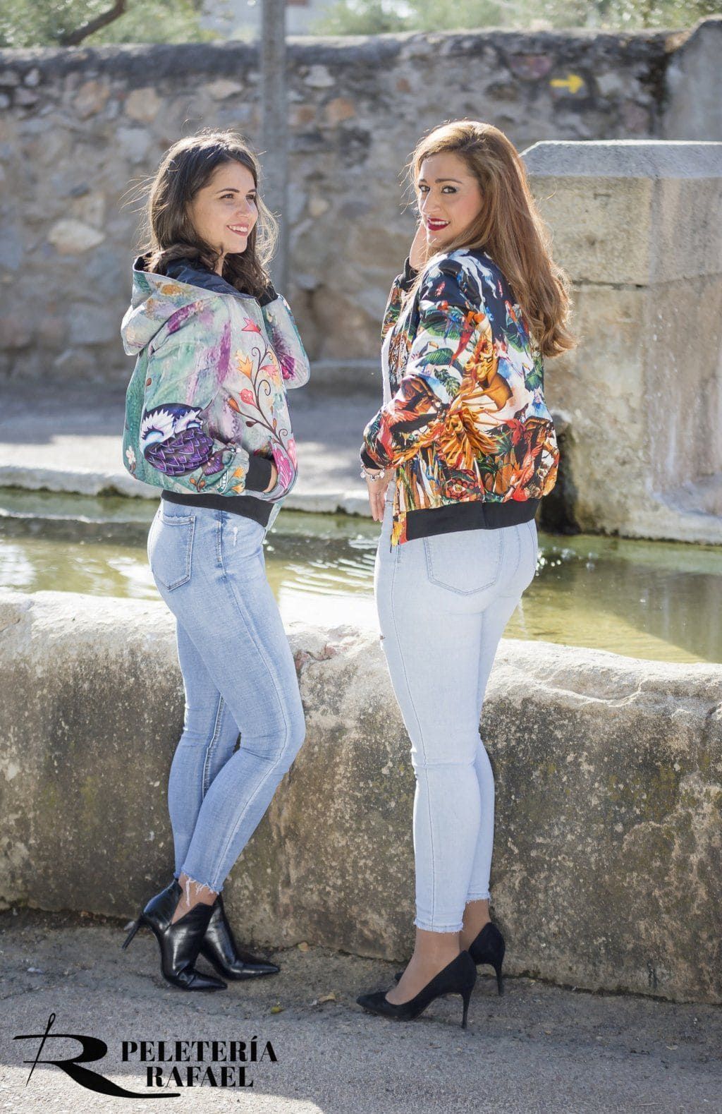 Dos mujeres con chaquetas estampadas y jeans posando al aire libre junto a una estructura de piedra.