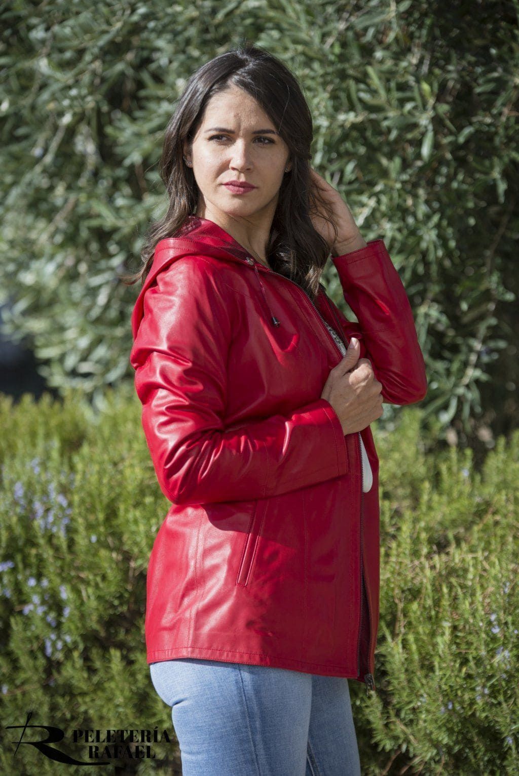 Mujer con impermeable rojo, jeans claros, parada afuera, mirando a la cámara y con la mano tocando su cabello.