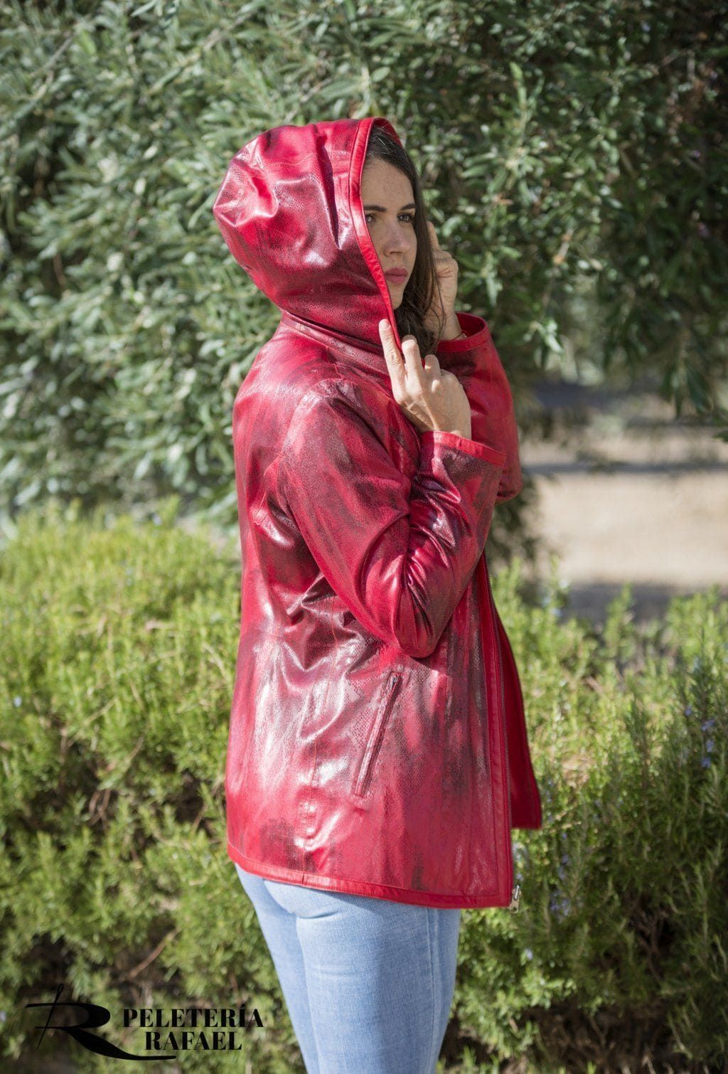 Mujer con impermeable con capucha roja, parada afuera junto a arbustos verdes.