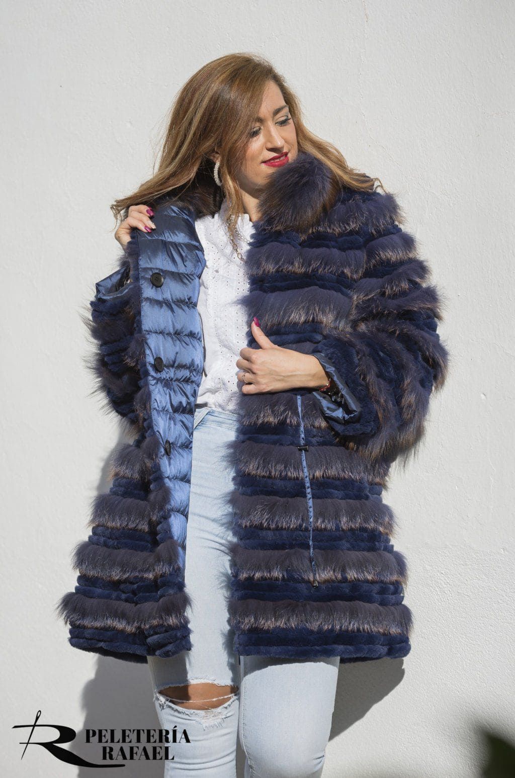 Mujer con abrigo de piel azul marino, camisa blanca y vaqueros rotos. Apoyada en una pared blanca.