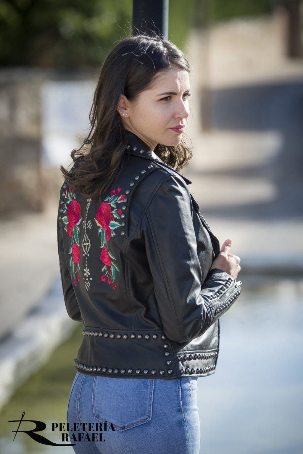 Mujer con chaqueta de cuero negra con bordado de flores rojas, de pie al aire libre, mirando hacia otro lado.