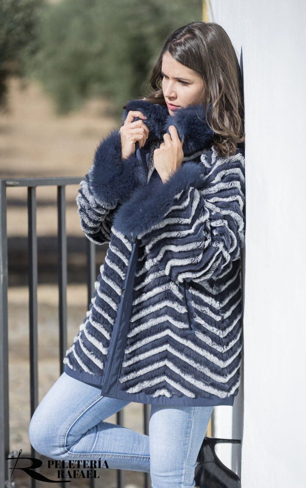 Mujer con una chaqueta azul ribeteada de piel, apoyada contra una pared blanca y una barandilla de metal; entorno al aire libre.