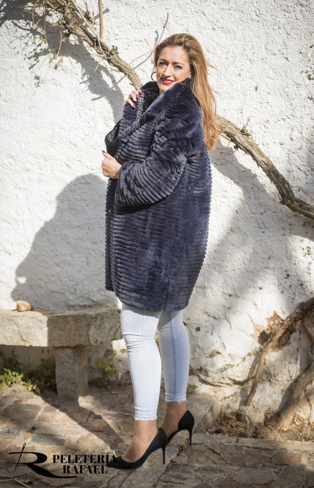 Una mujer con un abrigo azul marino, jeans claros y tacones está afuera, sonriendo.