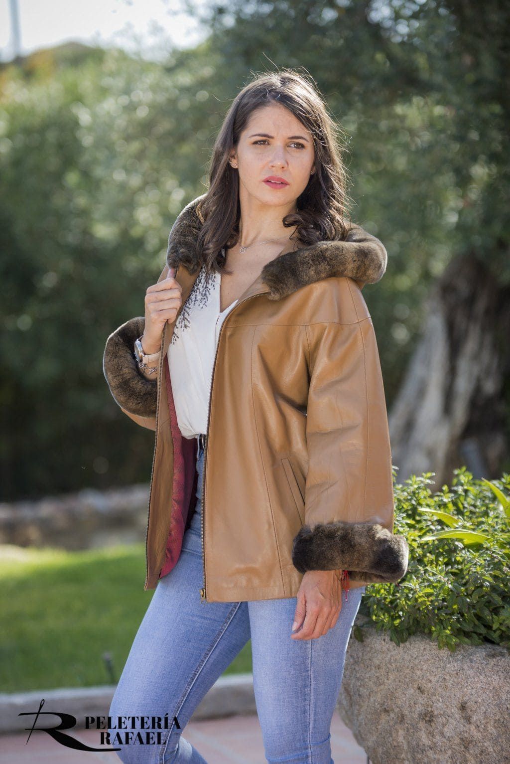 Mujer con abrigo color canela con ribete de piel, de pie al aire libre frente a la vegetación.