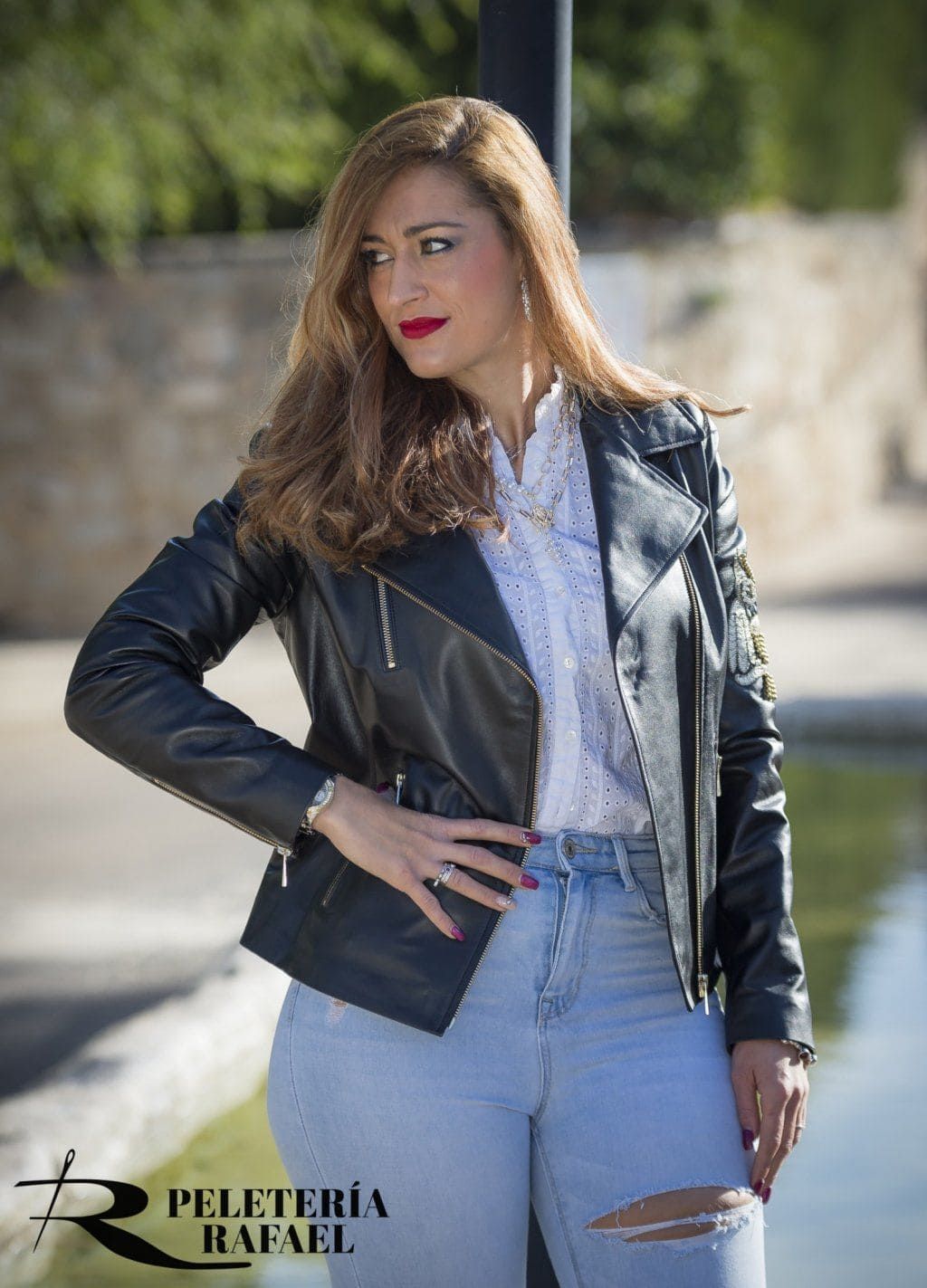 Mujer con chaqueta de cuero negra, jeans azul claro, mano en la cadera, mirando hacia un lado al aire libre.