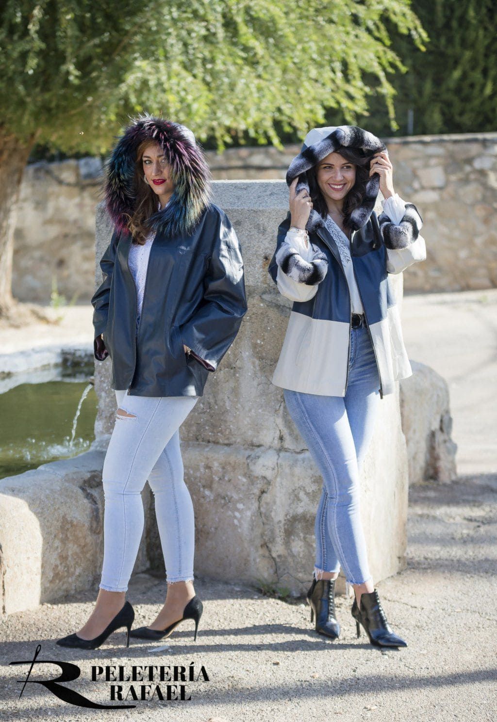 Dos mujeres vestidas con chaquetas forradas de piel, jeans y tacones posan al aire libre cerca de una fuente.