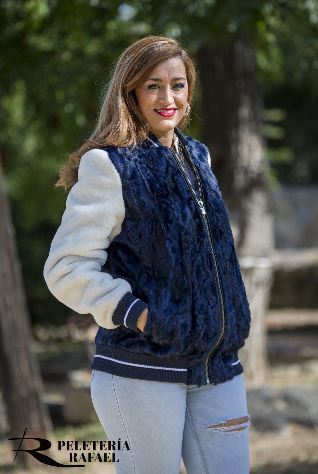 Mujer con una chaqueta bomber de piel azul y blanca y jeans de lavado claro, sonriendo al aire libre.