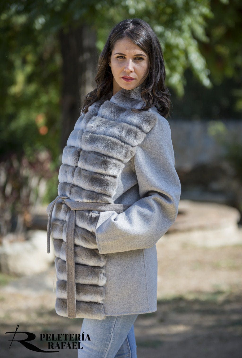 Mujer con abrigo gris con ribete de piel, de pie al aire libre. Lleva vaqueros azules, con las manos en los bolsillos, mirando a la cámara.
