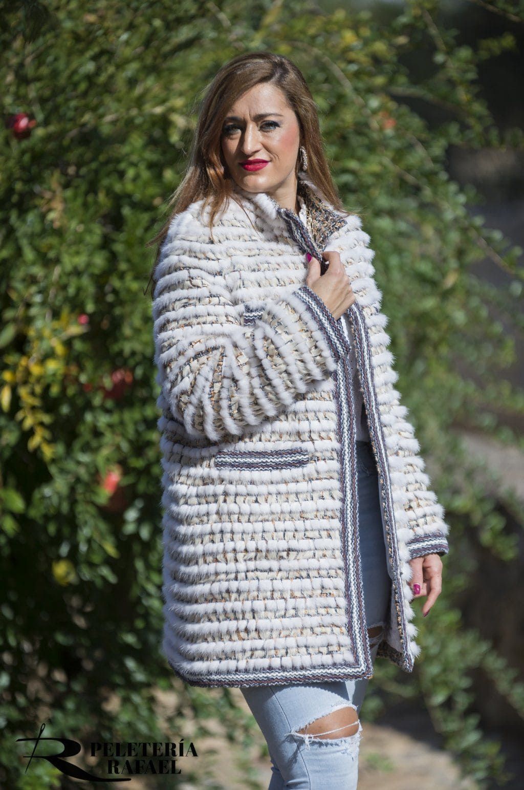 Mujer que viste un abrigo blanco texturizado con ribetes grises, de pie al aire libre cerca de un follaje verde, sonriendo.