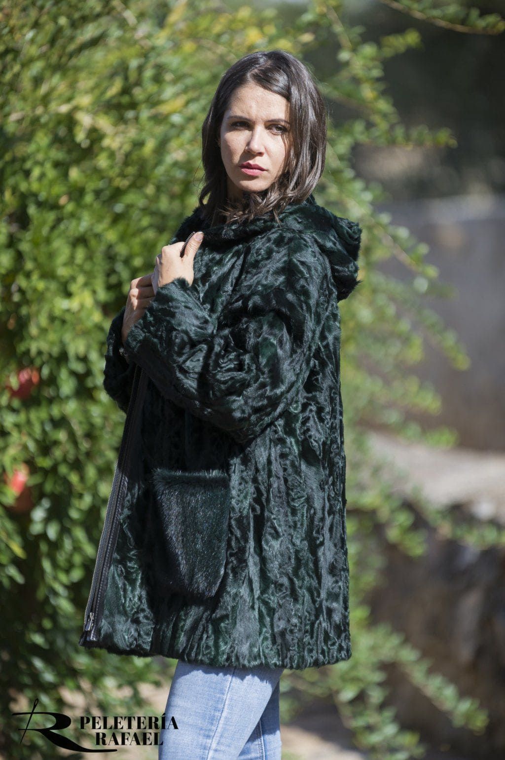 Mujer con un abrigo de piel verde oscuro con capucha y bolsillos, vistiendo jeans azul claro, parada afuera.