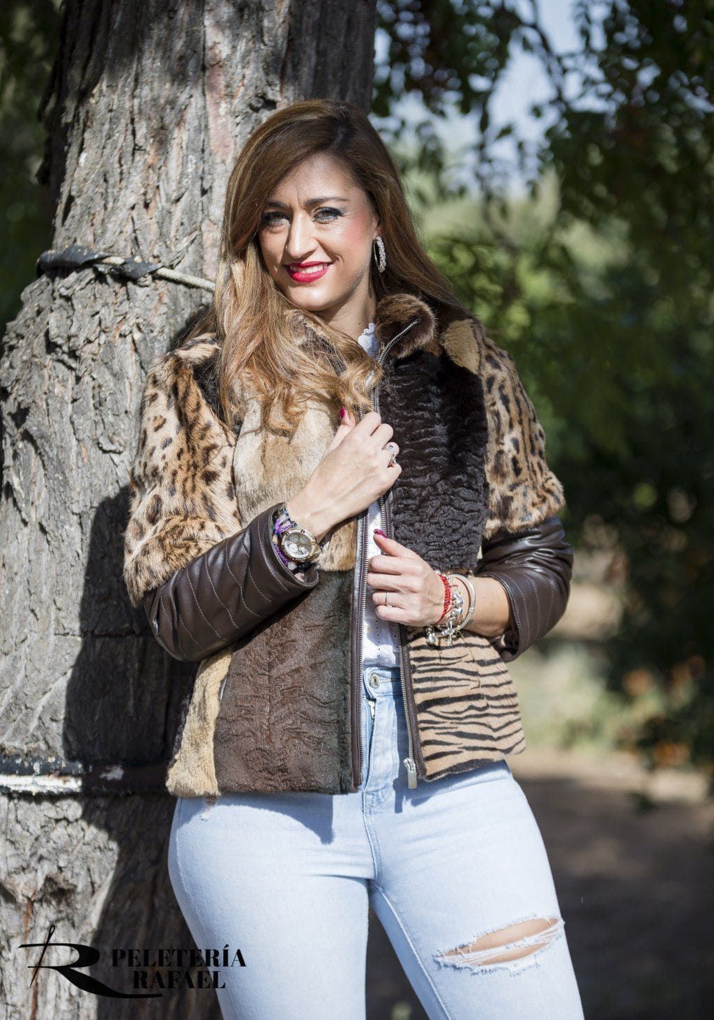 Una mujer con chaqueta ribeteada de piel y vaqueros claros posa junto a un árbol. Sonríe y toca la chaqueta.