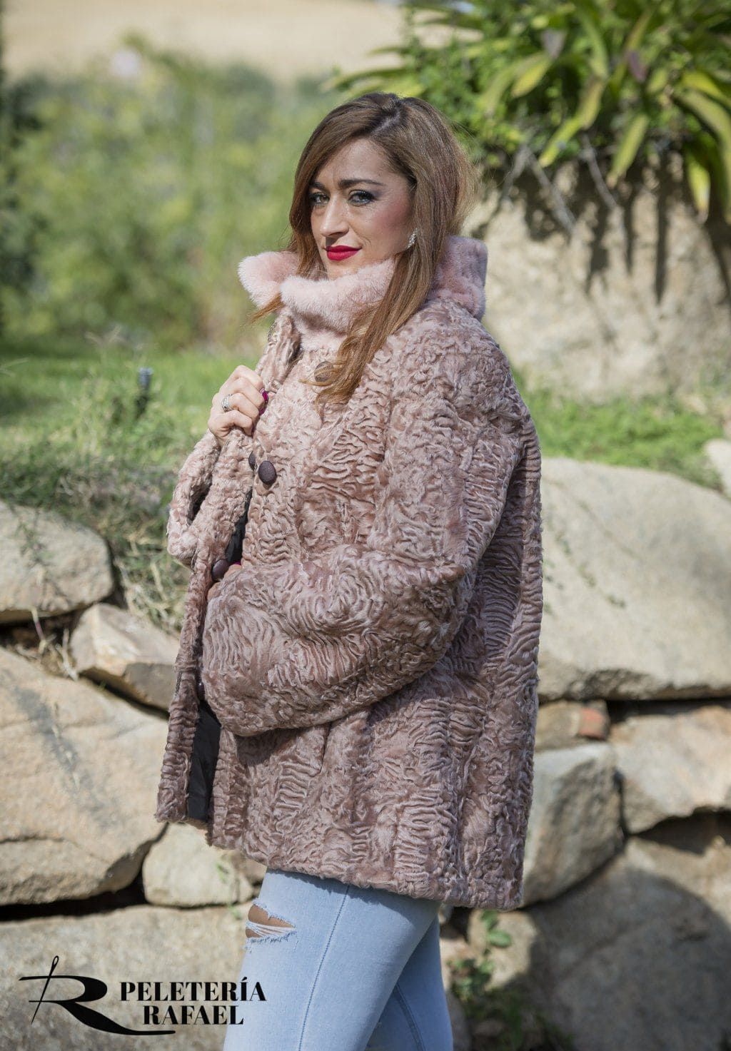 Mujer con un abrigo de piel rosa, jeans azul claro, lápiz labial rojo, parada junto a las rocas, al aire libre.
