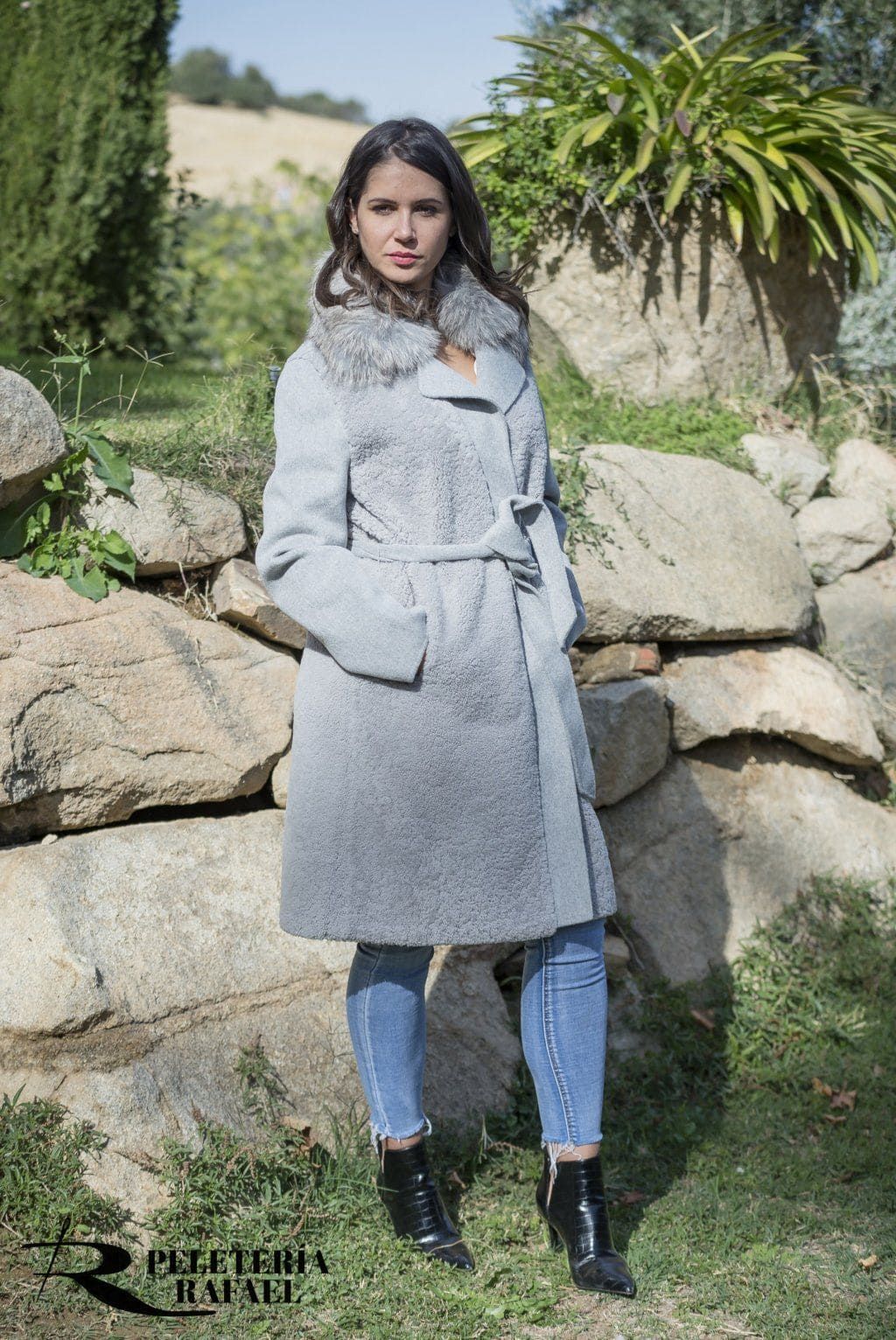 Mujer que lleva un abrigo gris con cuello de piel, abrochado con cinturón sobre vaqueros y botas, de pie al aire libre.