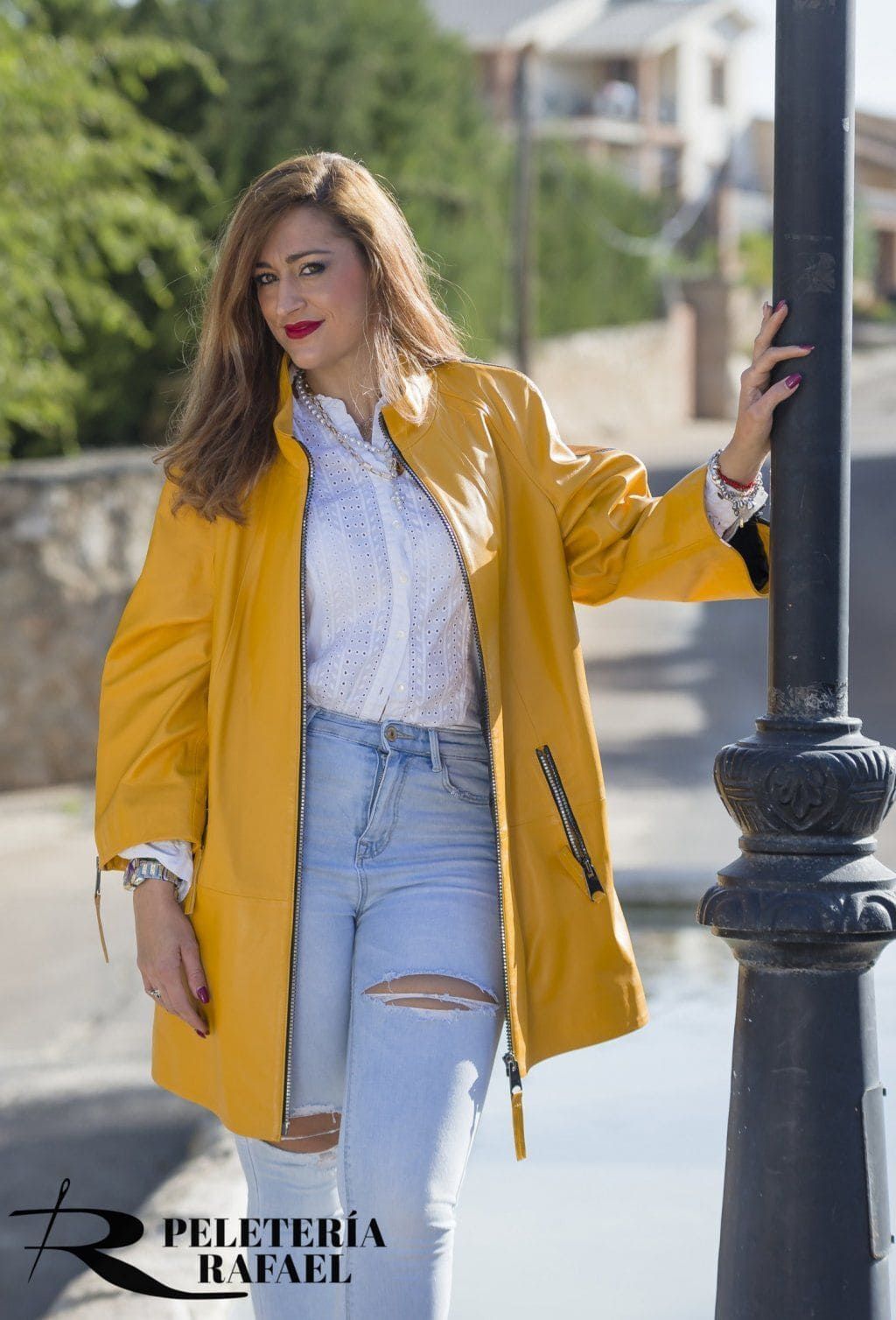 Mujer con abrigo amarillo, camisa blanca y jeans rotos, apoyada en un poste de luz al aire libre.