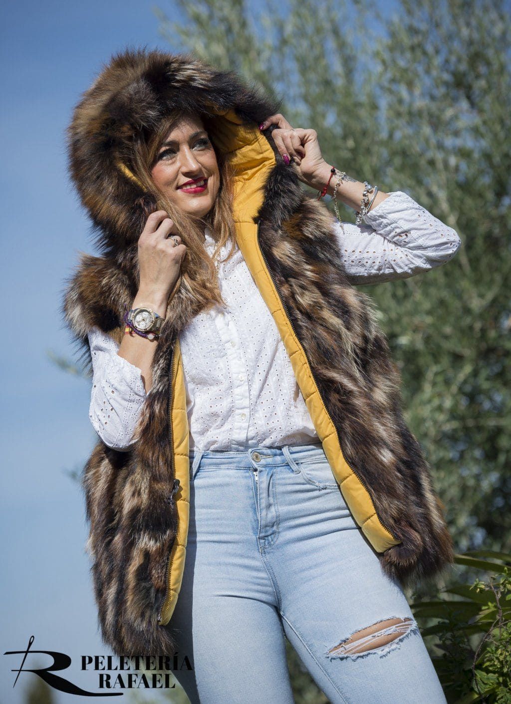 Mujer que lleva un chaleco de piel sintética con capucha forrada de amarillo y jeans rotos, sonriendo al aire libre.