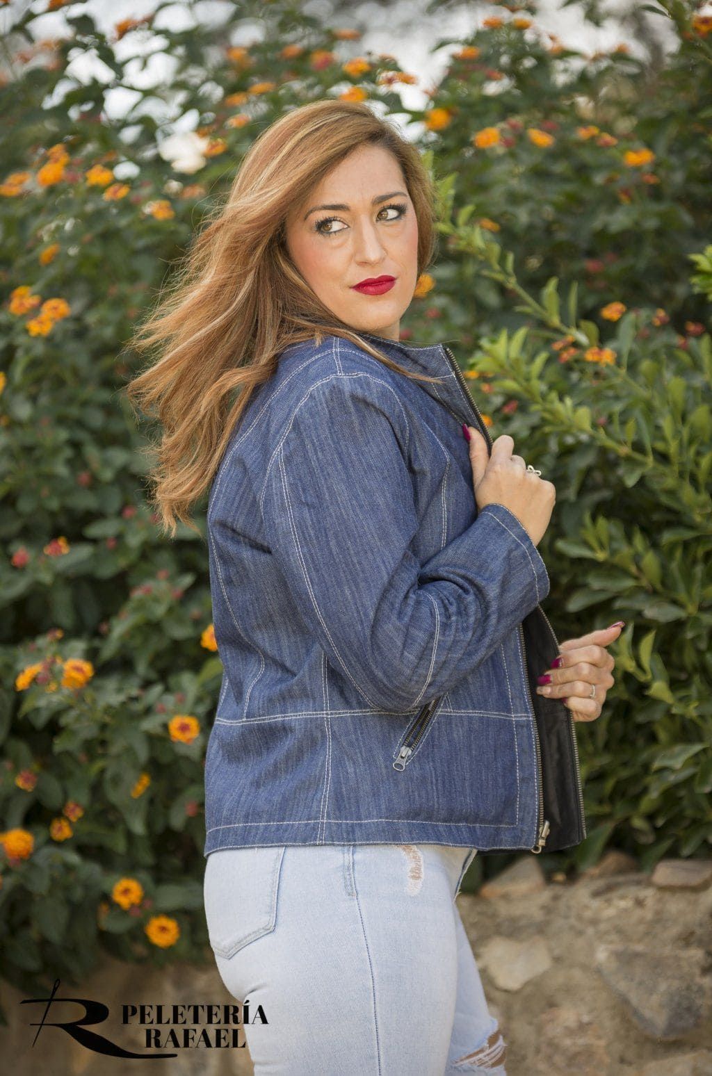 Mujer con chaqueta vaquera azul y pantalones vaqueros claros, posando al aire libre junto a arbustos en flor.