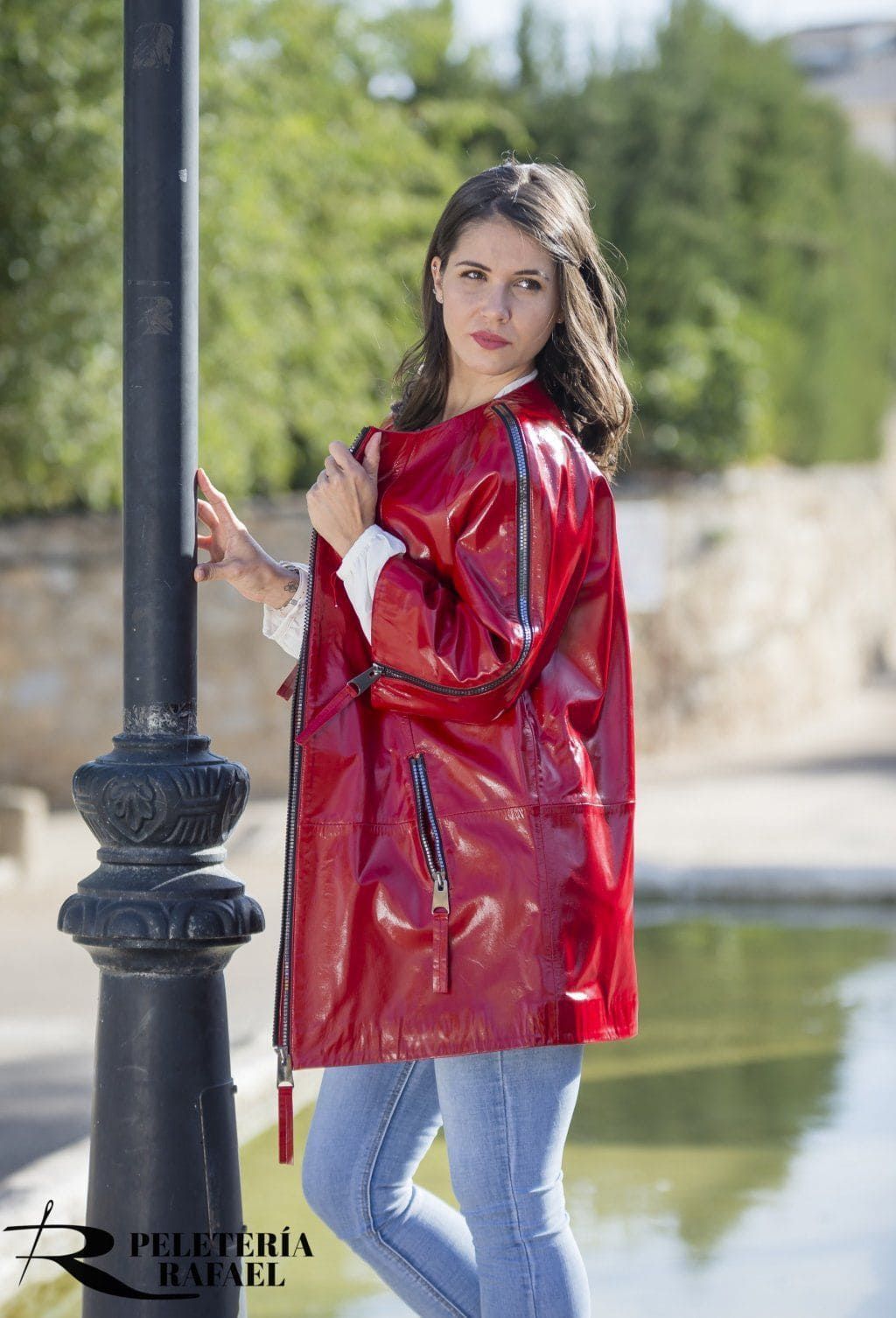 Mujer con chaqueta roja brillante y jeans junto a una farola al lado del agua.
