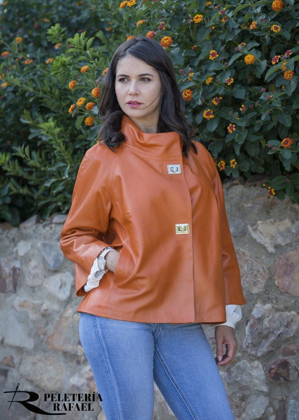 Mujer con chaqueta de cuero naranja, manos en los bolsillos, de pie al aire libre con follaje y pared de roca.