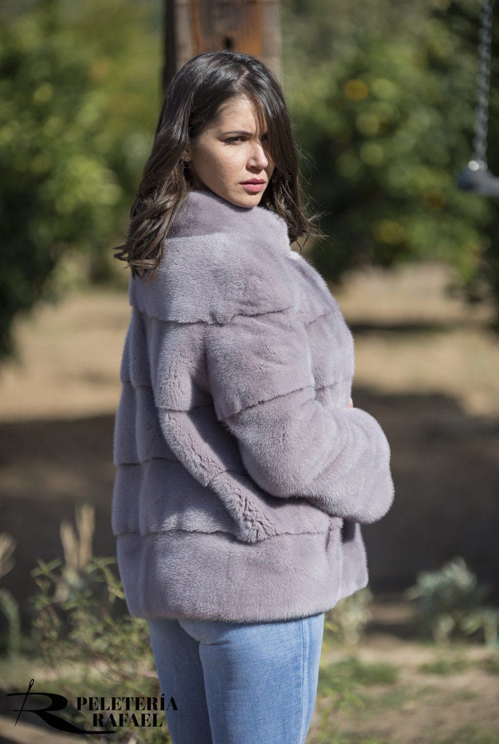Una mujer con un abrigo de piel de color violeta claro mira por encima del hombro, al aire libre.