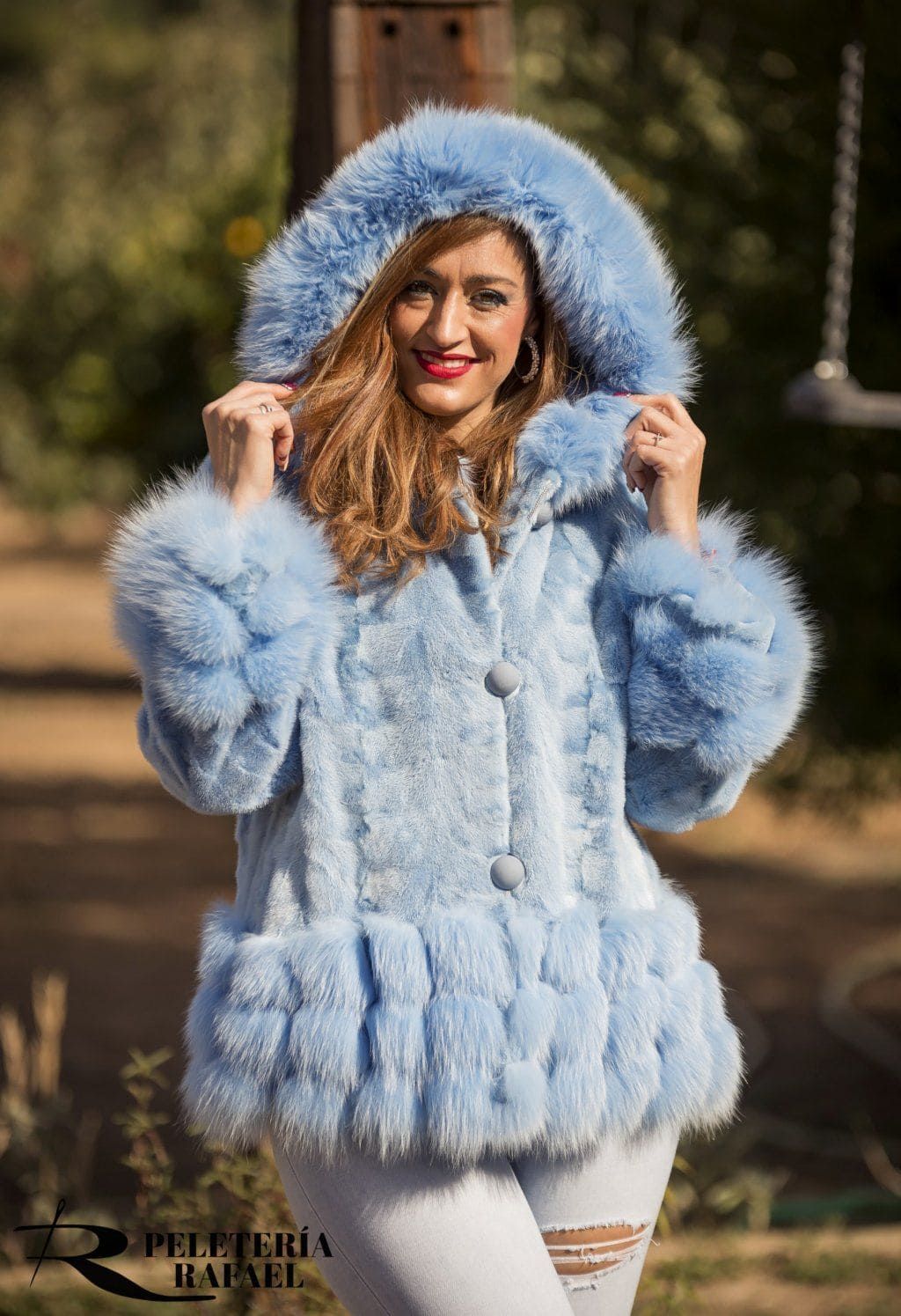 Mujer con un abrigo de piel azul claro con capucha y jeans blancos, sonriendo al aire libre.