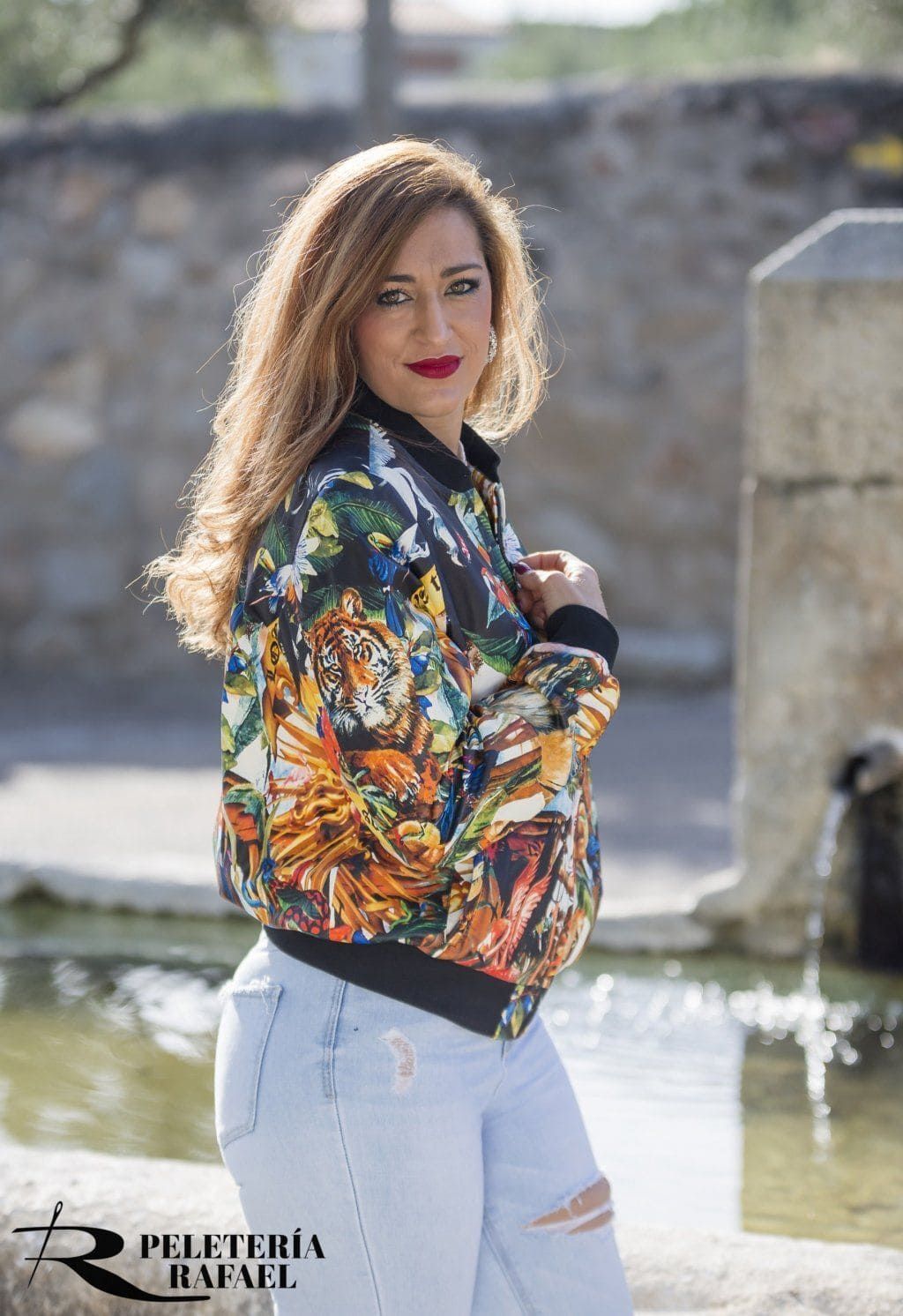 Una mujer con una chaqueta bomber estampada y unos vaqueros rotos está parada cerca de una fuente.