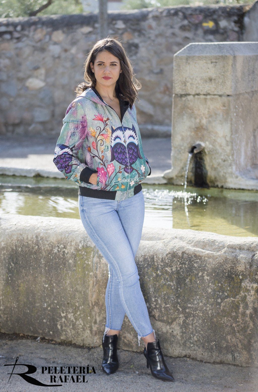 Una mujer con una chaqueta estampada y pantalones vaqueros está junto a una fuente de piedra.