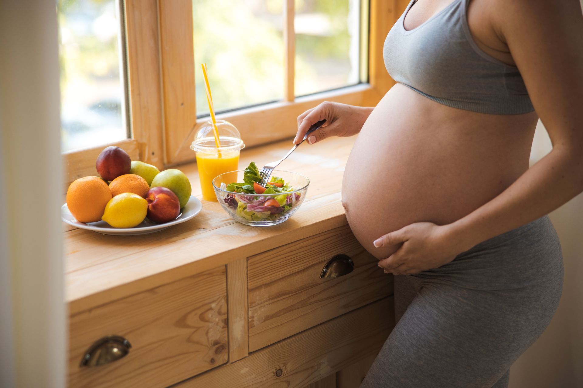 Une femme enceinte en tenue de sport grise mange de la salade et boit des fruits et du jus près d'une fenêtre.