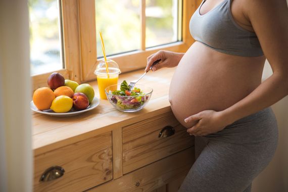 Une femme enceinte en tenue de sport grise mange de la salade et boit des fruits et du jus près d'une fenêtre.