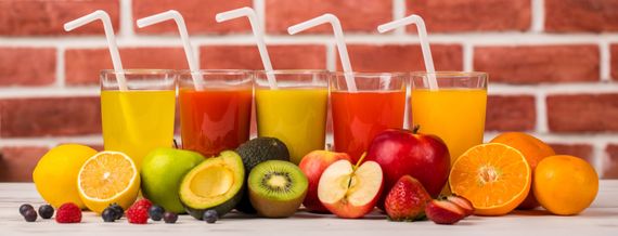Des verres de jus colorés avec des pailles sont placés devant des fruits contre un mur de briques.
