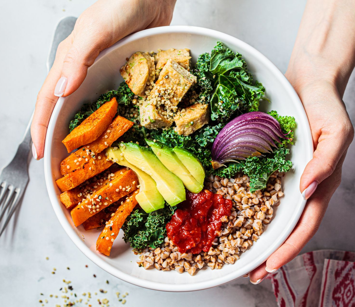 Mains tenant un bol blanc rempli de nourriture végétalienne colorée : patate douce, tofu, chou frisé, avocat, oignon rouge et sauce.