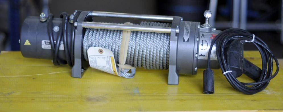 Un treuil argenté avec un câble en acier enroulé sur une surface jaune, avec une télécommande et des fils noirs.