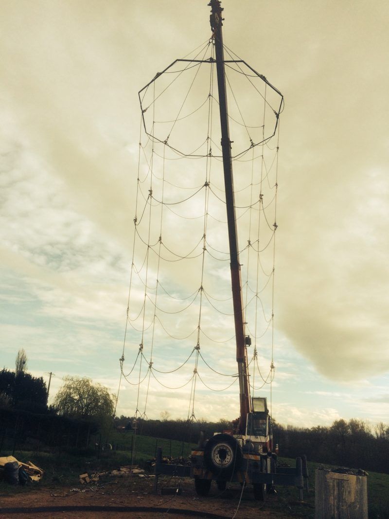 Une grue soulève une grande structure en filet. Un ciel nuageux forme l'arrière-plan.