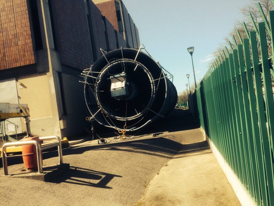 Grande structure cylindrique sur un chemin en béton, à côté d'un bâtiment et d'une clôture verte.