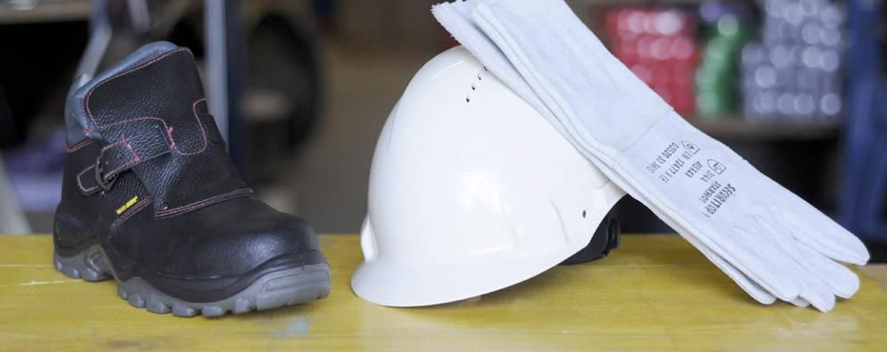 Bottes de travail noires, casque de chantier blanc et longs gants de travail blancs sur une surface jaune.