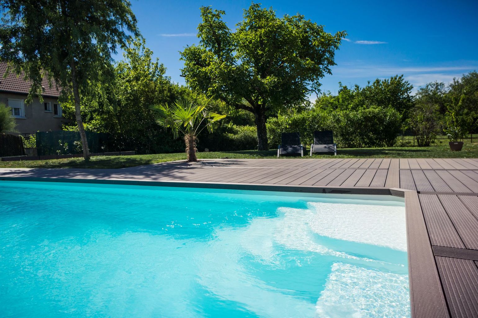 Piscine avec marches, eau turquoise, terrasse en bois, arbres, ciel bleu.