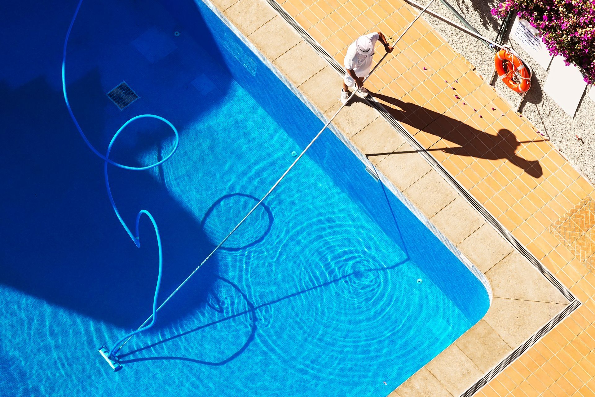 Personne passant l'aspirateur dans une piscine bleue avec un long tuyau, à côté de carrelage beige.