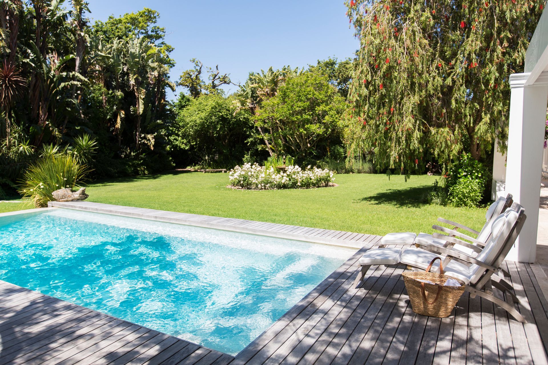 Piscine et terrasse en bois entourées d'herbe verte et d'arbres par une journée ensoleillée.