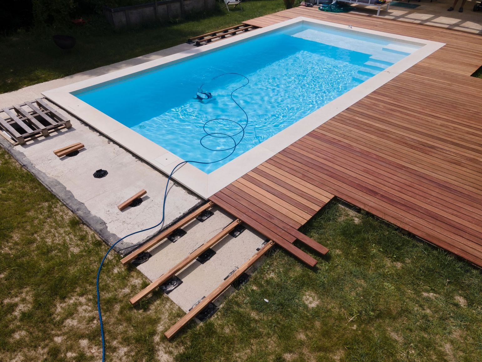 Piscine avec terrasse en bois en construction sur une pelouse. On aperçoit les matériaux de construction et un robot nettoyeur.
