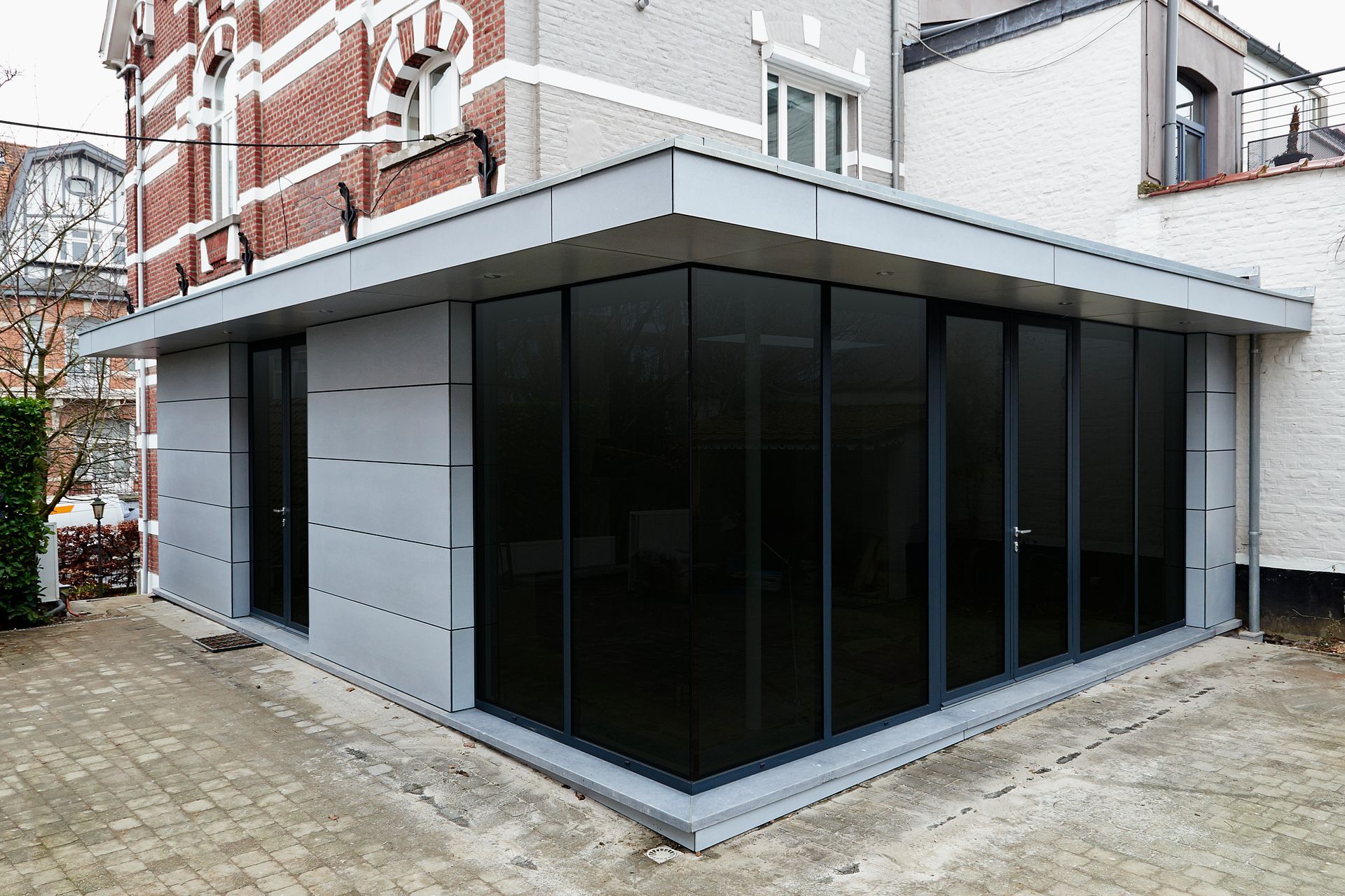 Extension moderne revêtue de gris, avec des fenêtres teintées foncées, attenante à un bâtiment en briques.