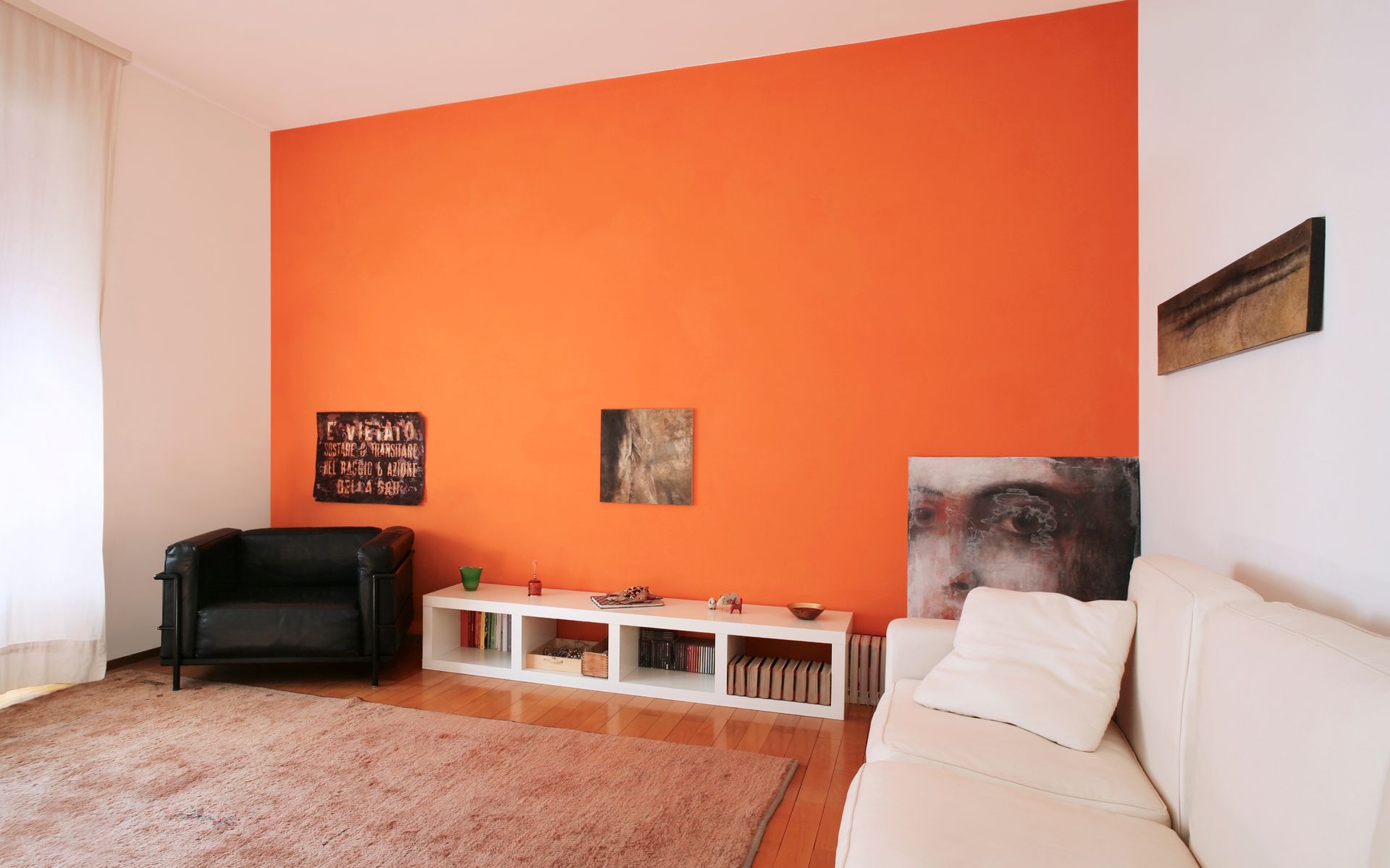 Salon avec un mur d'accent orange, un fauteuil en cuir noir, un canapé blanc et des étagères en bois.