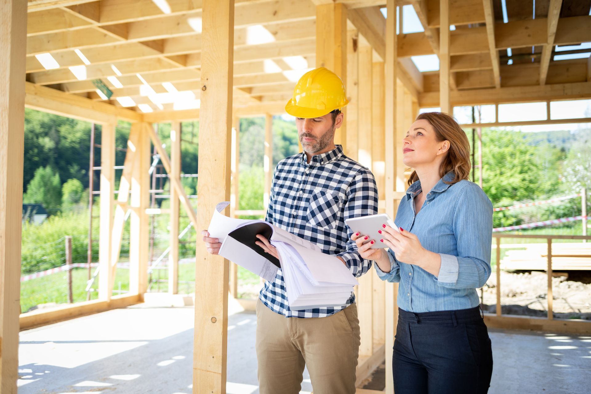 Chantier : un homme portant un casque de chantier et une femme examinent des plans ; la charpente en bois est visible.